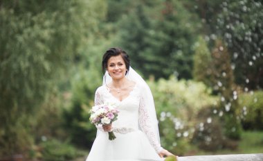 Zarif beyaz elbiseli güzel esmer gelin elinde buket ile güzel ağaçlar poz veriyor.