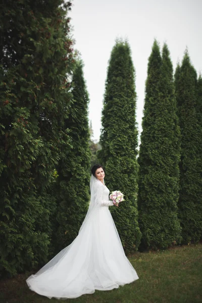 Zarif beyaz elbiseli güzel esmer gelin elinde buket ile güzel ağaçlar poz veriyor.