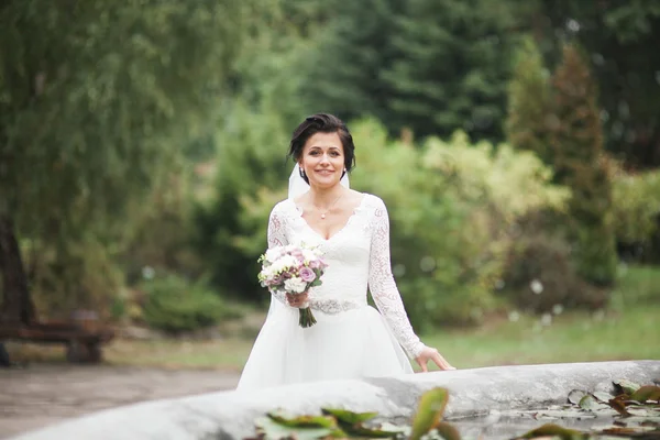 Bela noiva morena em vestido branco elegante segurando buquê posando árvores limpas — Fotografia de Stock