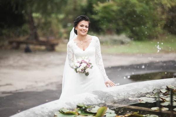 Beautiful brunette bride in elegant white dress holding bouquet posing neat trees — Stock Photo, Image