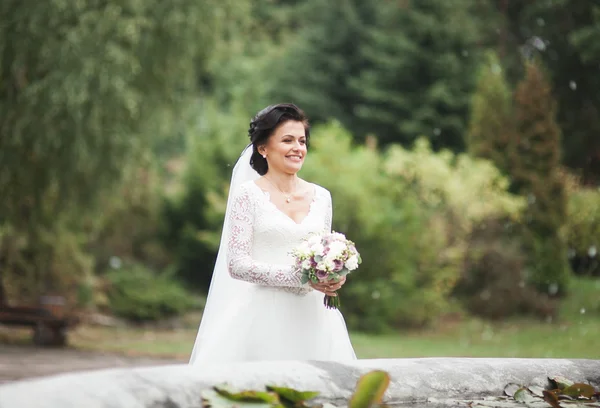 Bela noiva morena em vestido branco elegante segurando buquê posando árvores limpas — Fotografia de Stock