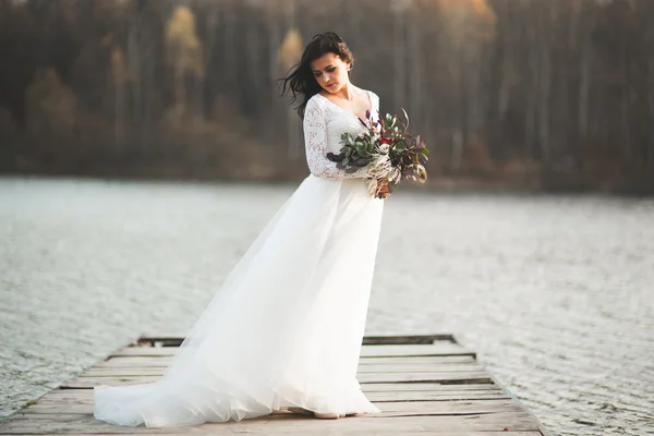 Superbe mariée brune en robe élégante tenant bouquet posant près de la forêt et du lac — Photo