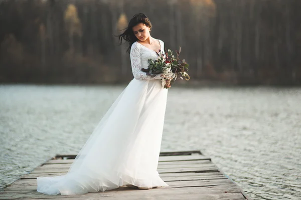 Prachtige brunette bruid in elegante jurk bedrijf boeket poseren in de buurt van bos en meer — Stockfoto