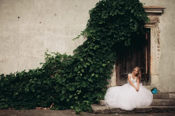 Krásná mladá blonďatá nevěsta s svatební kytice sedí na schodech pozadí nádherné rostliny — Stock fotografie