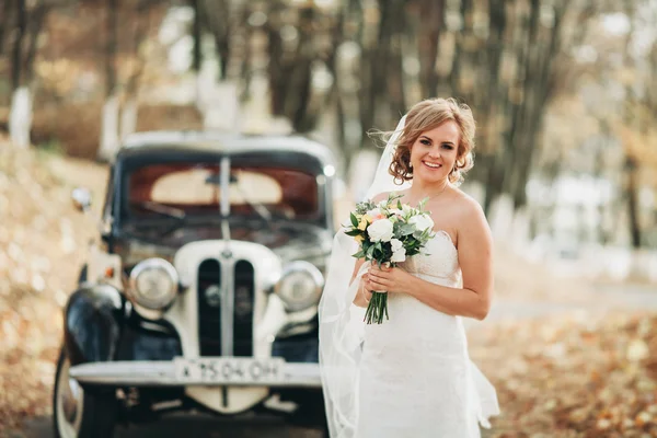 Mooie gelukkige bruid met boeket in de buurt van retro auto in de herfst — Stockfoto
