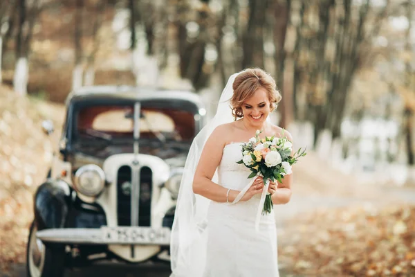 Mooie gelukkige bruid met boeket in de buurt van retro auto in de herfst — Stockfoto