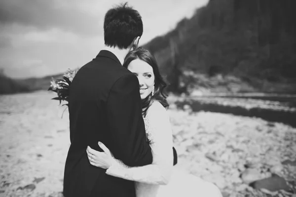 Happy bröllopsparet, bruden och brudgummen poserar snyggt flod mot bakgrund av bergen — Stockfoto