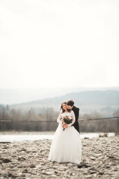 Happy wedding couple, bride and groom posing neat river against backdrop of the mountains — Stock Photo, Image