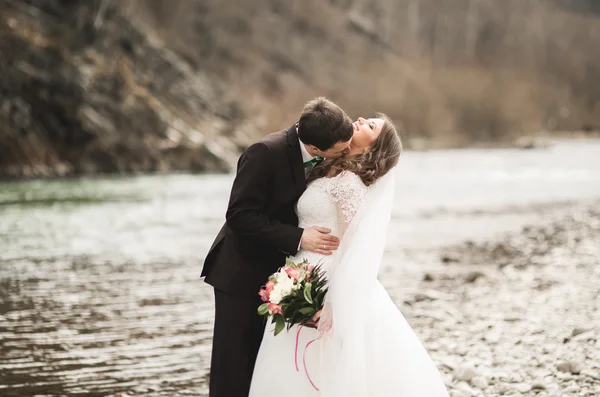 Feliz pareja de boda, novia y novio posando río aseado contra el telón de fondo de las montañas —  Fotos de Stock