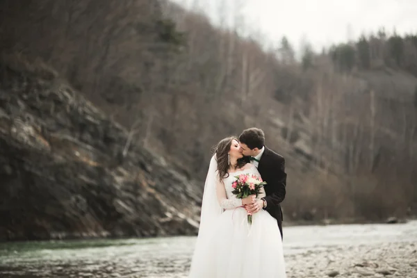 Felice coppia di nozze, sposa e sposo posa fiume pulito sullo sfondo delle montagne — Foto Stock