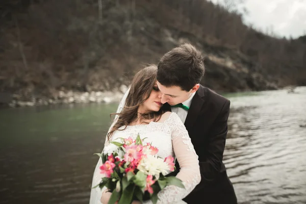 Pasangan pengantin yang cantik berciuman dan berpelukan di dekat sungai dengan batu — Stok Foto