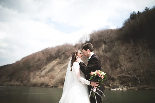 Feliz pareja de boda, novia y novio posando río aseado contra el telón de fondo de las montañas — Foto de Stock