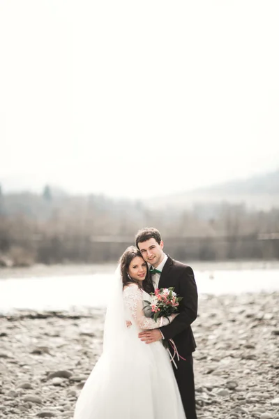 Красивая свадебная пара целуется и обнимается у реки с камнями — стоковое фото