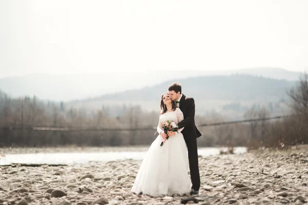 Feliz pareja de boda, novia y novio posando río aseado contra el telón de fondo de las montañas — Foto de Stock