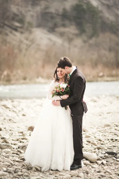 Felice coppia di nozze, sposa e sposo posa fiume pulito sullo sfondo delle montagne — Foto Stock