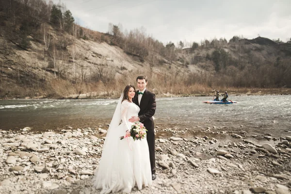 Красивая свадебная пара целуется и обнимается у реки с камнями — стоковое фото