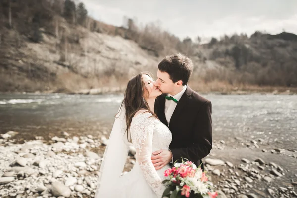 Feliz pareja de boda, novia y novio posando río aseado contra el telón de fondo de las montañas —  Fotos de Stock