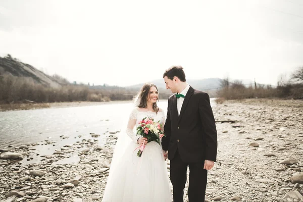 Felice coppia di nozze, sposa e sposo posa fiume pulito sullo sfondo delle montagne — Foto Stock