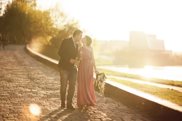 Bröllop par, rykta och klä poserar nära floden med ett glas vid solnedgången — Stockfoto