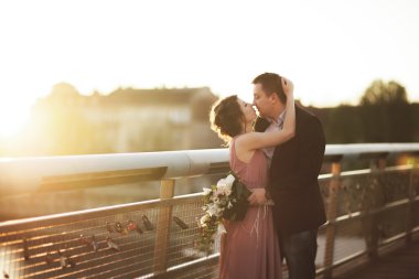 Şık Düğün çifti, damat, Gelin öpüşme ve günbatımında bir köprüde sarılma pembe elbise ile sevgi dolu