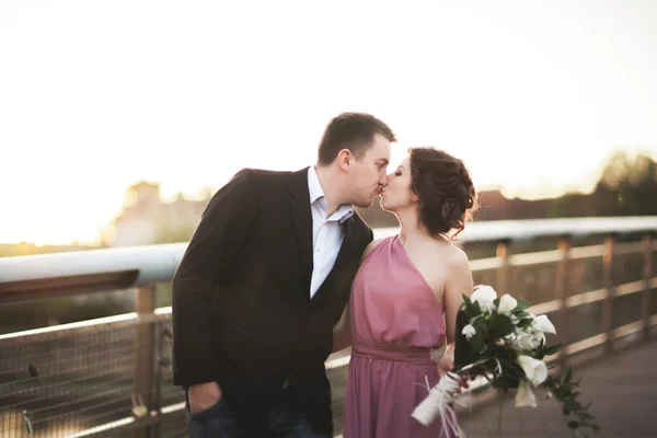 Stylish loving wedding couple, groom, bride with pink dress kissing and hugging on a bridge at sunset — Stock Photo, Image