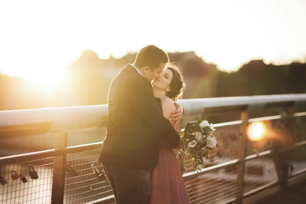 Stilvoll liebende Hochzeitspaar, Bräutigam, Braut mit rosa Kleid küsst und umarmt auf einer Brücke bei Sonnenuntergang — Stockfoto