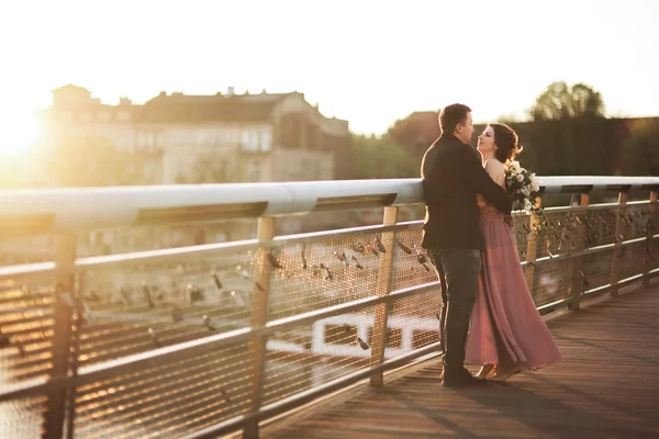 Stylový, milující Svatební pár, ženich, nevěsta s růžové šaty, líbání a objímání při západu slunce na mostě — Stock fotografie