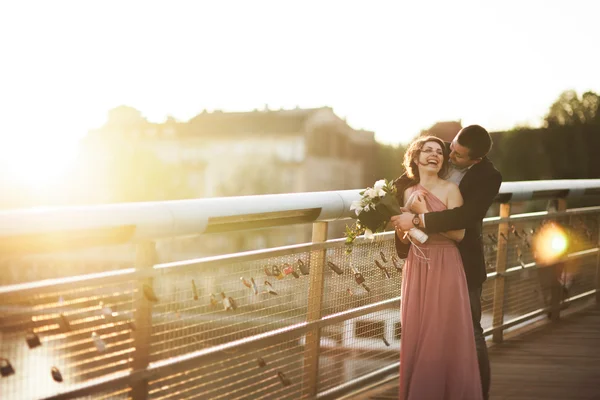 Elegante pareja de boda amorosa, novio, novia con vestido rosa besándose y abrazándose en un puente al atardecer — Foto de Stock
