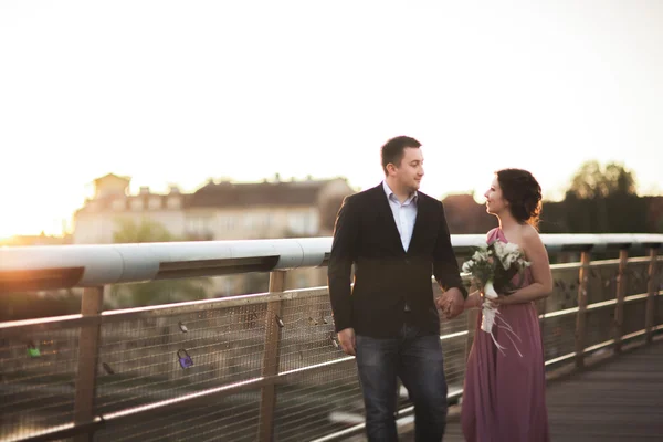 Snygg kärleksfull bröllopsparet, brudgummen, brud med rosa klänning kyssas och kramas på en bro vid solnedgången — Stockfoto