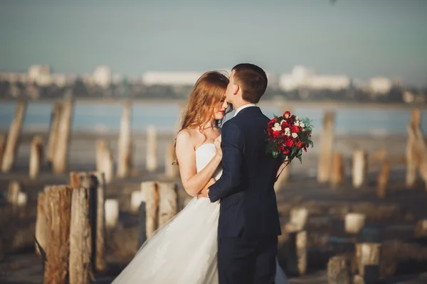 Beautiful young wedding couple, bride and groom posing near wooden poles on the background sea — Stock Photo, Image