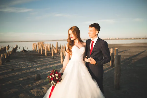 Pareja de boda, novio, novia con ramo posando cerca del mar al atardecer — Foto de Stock