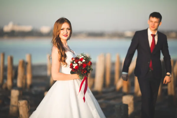Красива молода весільна пара, наречена і наречений позує біля дерев'яних полюсів на фоні моря — стокове фото