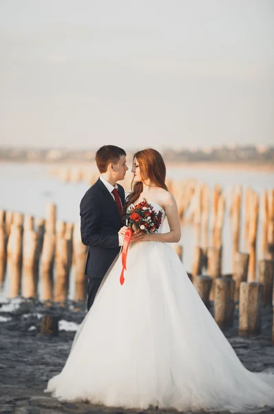 Casal de casamento jovem bonito, noiva e noivo posando perto de postes de madeira no fundo do mar — Fotografia de Stock