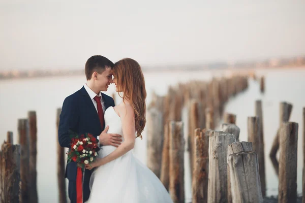 Casal de casamento, noivo, noiva com buquê posando perto do mar ao pôr do sol — Fotografia de Stock