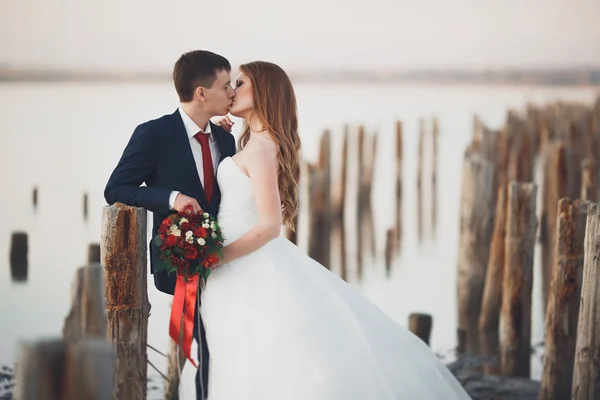 Beautiful young wedding couple, bride and groom posing near wooden poles on the background sea — Stock Photo, Image