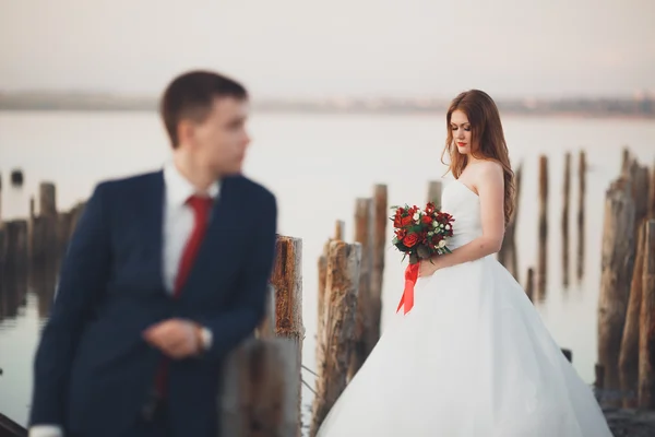 Beautiful young wedding couple, bride and groom posing near wooden poles on the background sea — Stock Photo, Image