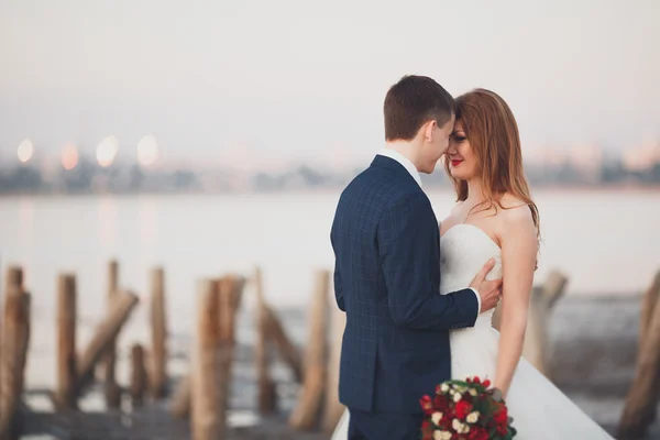 Wedding couple, groom, bride with bouquet posing near sea on sunset — Stock Photo, Image