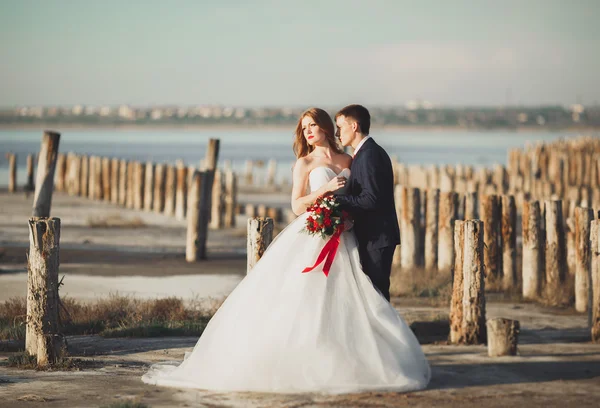 Hermosa joven pareja de boda, novia y novio posando cerca de postes de madera en el mar de fondo — Foto de Stock