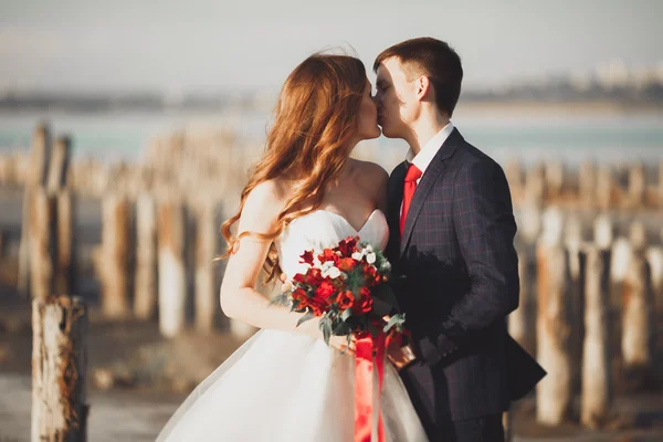 Весільна пара, наречений, наречена з букетом позує біля моря на заході сонця — стокове фото