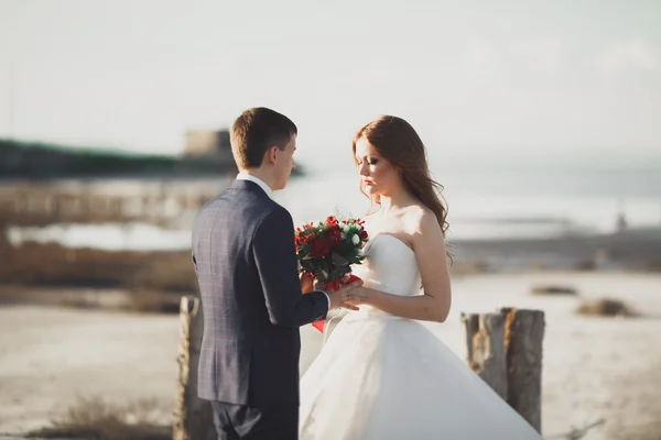 Casal de casamento, noivo, noiva com buquê posando perto do mar ao pôr do sol — Fotografia de Stock
