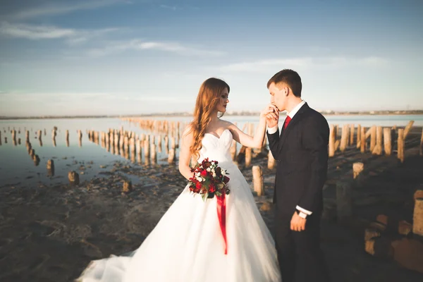 Casal de casamento, noivo, noiva com buquê posando perto do mar ao pôr do sol — Fotografia de Stock