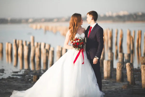 Hermosa joven pareja de boda, novia y novio posando cerca de postes de madera en el mar de fondo — Foto de Stock