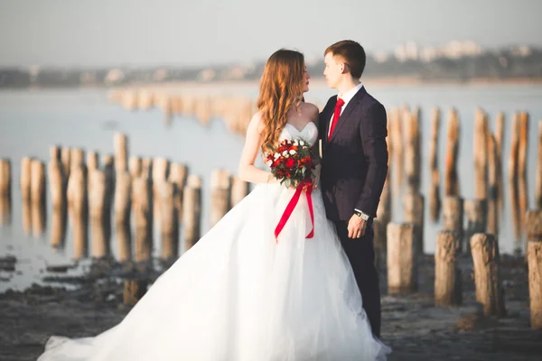 Casal de casamento jovem bonito, noiva e noivo posando perto de postes de madeira no fundo do mar — Fotografia de Stock