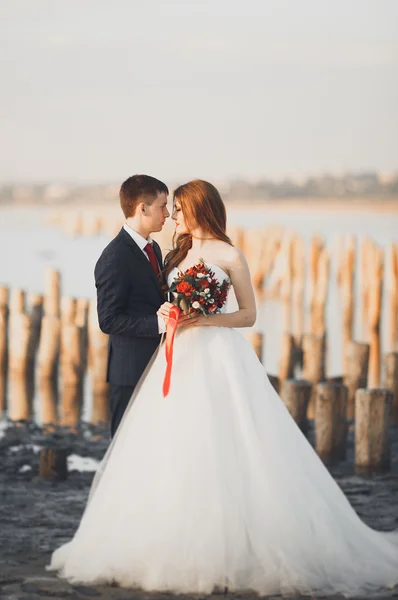 Bella giovane coppia di nozze, sposa e sposo in posa vicino pali di legno sullo sfondo mare — Foto Stock