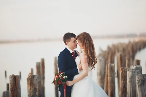 Casal de casamento jovem bonito, noiva e noivo posando perto de postes de madeira no fundo do mar — Fotografia de Stock