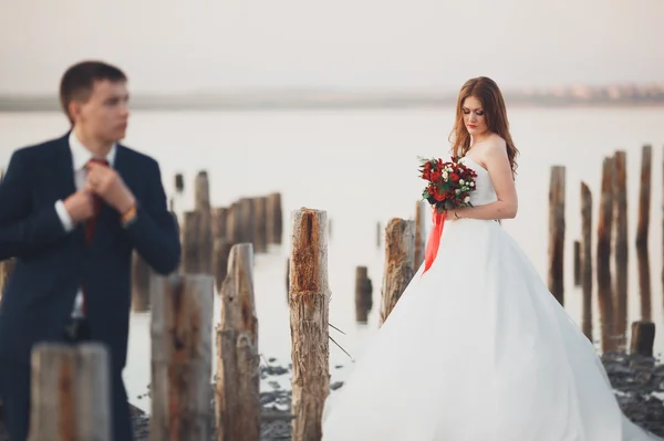 Schöne junge Hochzeitspaar, Braut und Bräutigam posiert in der Nähe von Holzpfählen auf dem Hintergrund Meer — Stockfoto