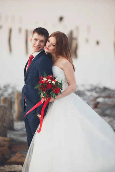 Свадебная пара, жених, невеста с букетом, позирующим у моря на закате — стоковое фото