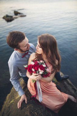 Düğün çifti mavi denizin yanındaki kayalarda öpüşüyor ve sarılıyor.