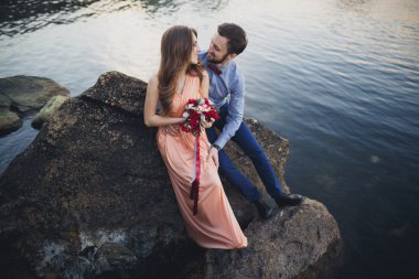 Düğün çifti mavi denizin yanındaki kayalarda öpüşüyor ve sarılıyor.