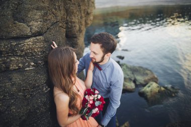 Düğün çifti mavi denizin yanındaki kayalarda öpüşüyor ve sarılıyor.